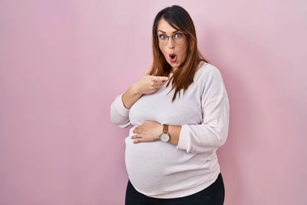妊娠中の女性はピンクの背景に立って驚いて指で側面を指して 口を開けて驚いて表情 — ストック写真