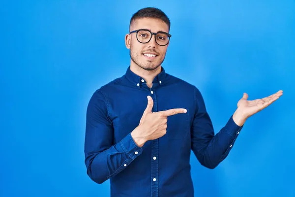 Joven Hombre Hispano Con Gafas Sobre Fondo Azul Asombrado Sonriendo —  Fotos de Stock