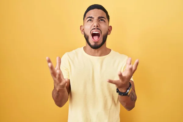 黄色の背景に立っている若いヒスパニック系の男は狂気と怒りの叫びと積極的な表現と腕を上げて叫んでいた フラストレーションコンセプト — ストック写真