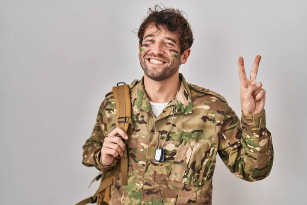 ヒスパニック系の若い男が迷彩軍の制服を着てカメラを見て笑みを浮かべて勝利のサインを行う指を示しています — ストック写真