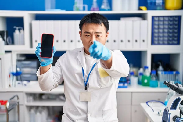 Young chinese man working at scientist laboratory holding smartphone pointing with finger to the camera and to you, confident gesture looking serious