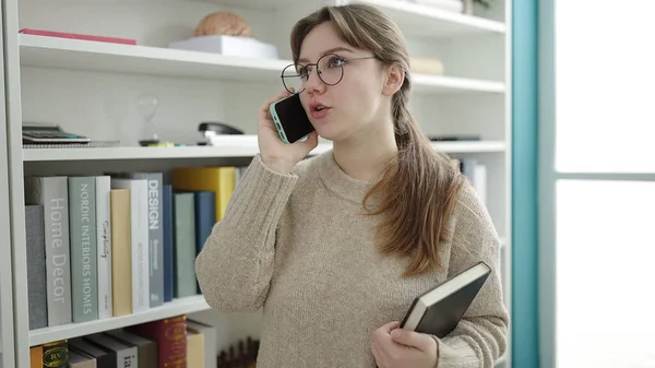 若いですブロンド女性学生話して上のスマートフォン保持本で図書館大学 — ストック写真