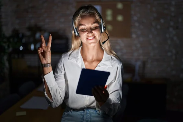 Young blonde woman working at the office at night smiling with happy face winking at the camera doing victory sign with fingers. number two.