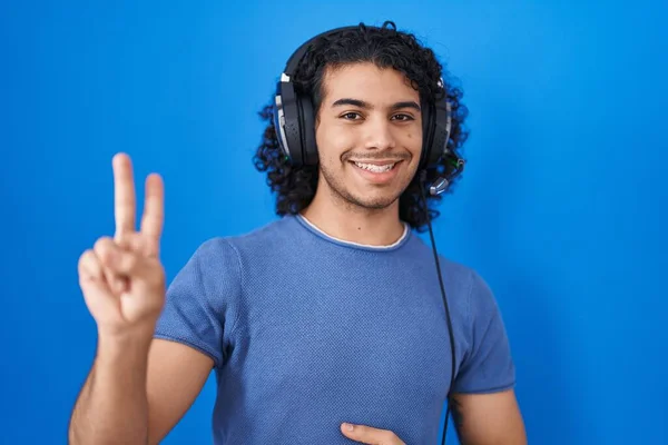ヘッドフォンを使用して音楽に耳を傾け巻き毛を持つヒスパニック系の男は 勝利のサインを行う指を示すカメラを見て笑顔 — ストック写真