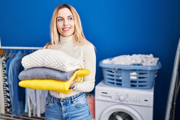 Young blonde woman smiling confident holding folded clothes at laundry room