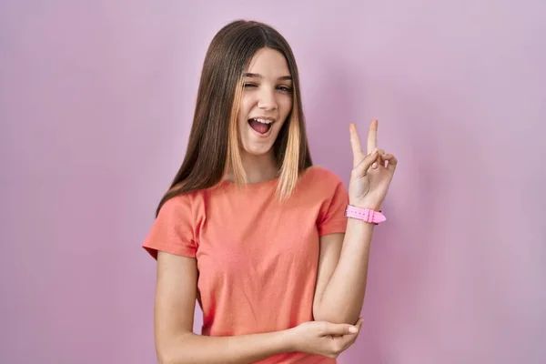 站在粉色背景上的少女高兴地微笑着 用手指在镜头前眨眼示意胜利 第二点 — 图库照片