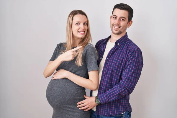 笑顔で明るい白い背景の上に立っている赤ちゃんを期待している若いカップル手と指で幸せと自然な表情で側面まで指して — ストック写真