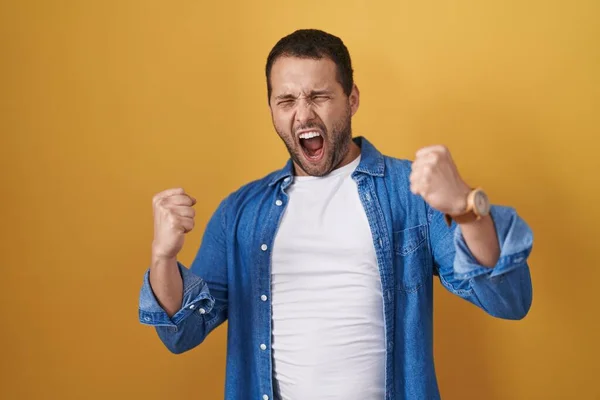 stock image Hispanic man standing over yellow background angry and mad raising fists frustrated and furious while shouting with anger. rage and aggressive concept. 