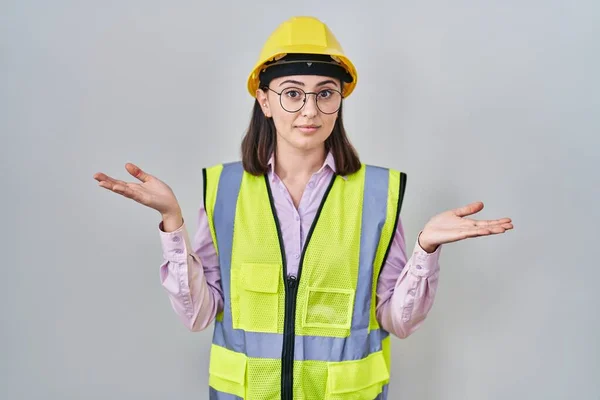 身穿建筑工人制服 头戴硬礼帽的西班牙裔女孩 满脸困惑 手臂和双手高举 怀疑概念 — 图库照片