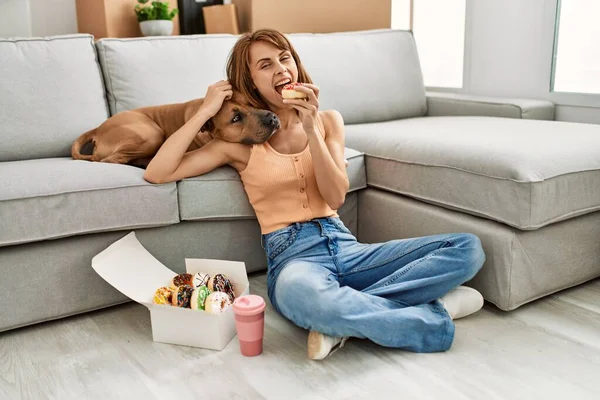 Young caucasian woman having breakfast sitting on floor with dog at home