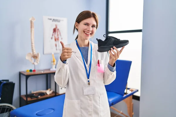 Joven Fisioterapeuta Mujer Sosteniendo Zapatillas Sonriendo Feliz Positivo Pulgar Hacia —  Fotos de Stock
