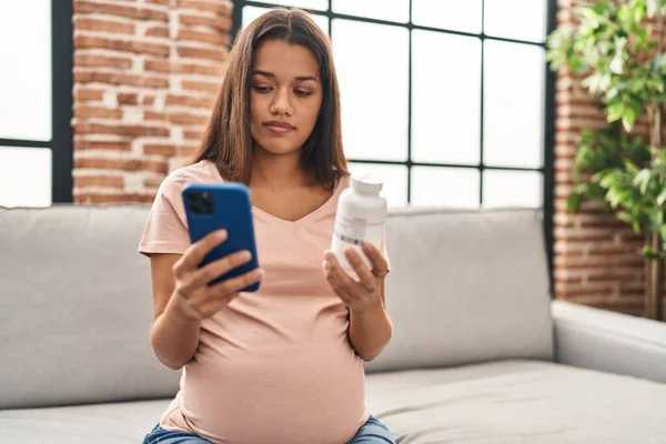 Młoda Latynoska Kobieta Ciąży Pomocą Smartfona Trzymając Butelkę Tabletek Domu — Zdjęcie stockowe