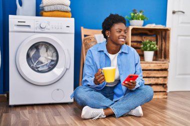 Akıllı telefon kullanan ve kahve içen Afrikalı Amerikalı kadın çamaşır odasında çamaşır makinesini bekliyor.
