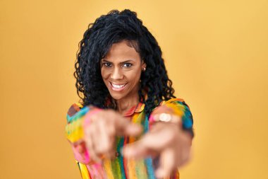 Middle age hispanic woman standing over yellow background pointing to you and the camera with fingers, smiling positive and cheerful 