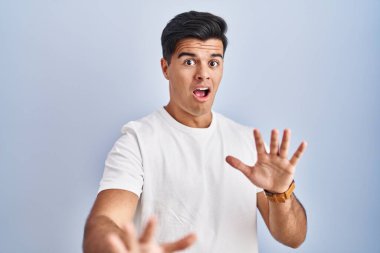 Hispanic man standing over blue background afraid and terrified with fear expression stop gesture with hands, shouting in shock. panic concept. 
