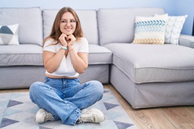 Oturma odasında yerde oturan genç, beyaz bir kadın. Gergin ve heyecanlı bir şekilde, elleri çenesinde yan tarafa bakıyor. 