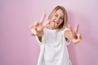 Pembe arka planda duran genç beyaz kadın iki elini de zafer işareti yaparak parmaklarını göstererek gülümsüyor. İki numara.. 