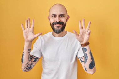 Young hispanic man with tattoos standing over yellow background showing and pointing up with fingers number nine while smiling confident and happy. 