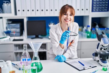 Young woman scientist smiling confident holding glasses at laboratory