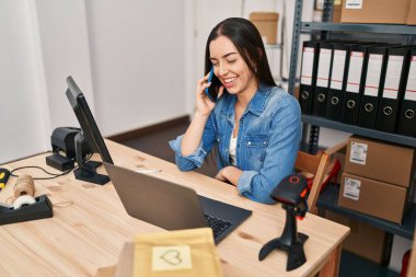 Young beautiful hispanic woman ecommerce business worker using laptop talking on smartphone at office