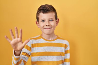 Sarı arka planda duran genç beyaz çocuk beş numarayı gösterip işaret ederken kendinden emin ve mutlu gülümsüyor.. 