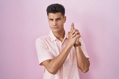Young hispanic man standing over pink background holding symbolic gun with hand gesture, playing killing shooting weapons, angry face 
