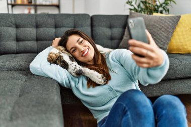 Evdeki akıllı telefonun yanında köpeğe sarılıp selfie çeken genç bir kadın.