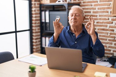 Gri saçlı yaşlı adam ofiste bilgisayar kullanarak çalışıyor rahatlıyor ve gözleri kapalı gülümsüyor parmaklarıyla meditasyon hareketi yapıyor. yoga konsepti. 