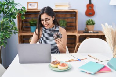 Young hispanic girl eating doughnut studying at home