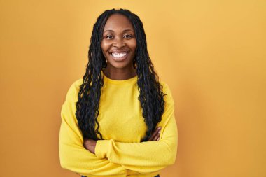 African woman standing over yellow background happy face smiling with crossed arms looking at the camera. positive person. 