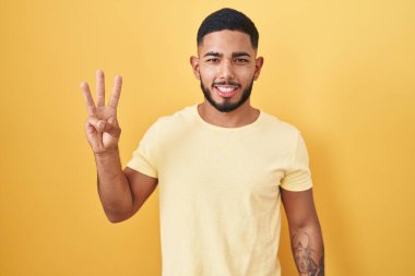 Young hispanic man standing over yellow background showing and pointing up with fingers number three while smiling confident and happy. 