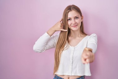 Young caucasian woman standing over pink background smiling doing talking on the telephone gesture and pointing to you. call me. 