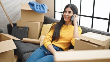 Genç, güzel, İspanyol bir kadın akıllı telefondan konuşuyor. Yeni evindeki kanepede oturuyor.