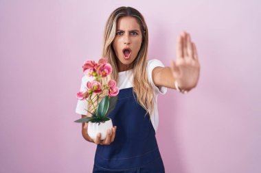 Sarışın, bahçıvan önlüğü giyen genç bir kadın elinde fabrikayla avuç içi hareketlerini durduruyor, kızgın ve hayal kırıklığı ifadesi takınıyor. 