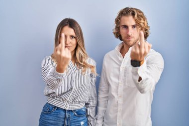 Young couple standing over blue background showing middle finger, impolite and rude fuck off expression 