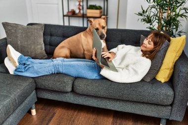 Genç beyaz kadın evdeki köpekle birlikte kanepede dizüstü bilgisayar kullanıyor.