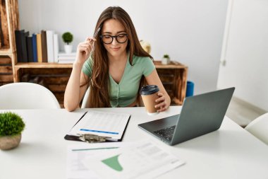 Young hispanic woman using laptop drinking coffee sitting on table at home