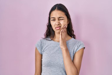 Genç Brezilyalı bir kadın pembe arka plan üzerine gündelik tişört giyip diş ağrısı ya da diş hastalığından dolayı acı dolu bir ifadeyle ağıza dokunuyor. dişçi 
