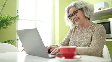 Middle age woman with grey hair using laptop sitting on table at home