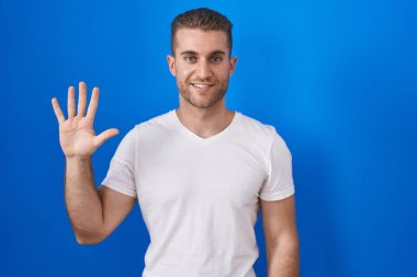 Mavi arka planda duran genç beyaz adam beş numarayı gösterip işaret ederken kendinden emin ve mutlu gülümsüyor.. 