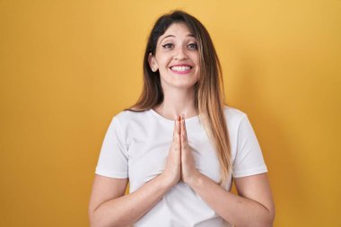 Sarı arka planda duran genç esmer kadın elleri birarada dua ediyor af diliyor kendinden emin bir şekilde gülümsüyor.. 