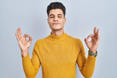 Young hispanic man standing over blue background relaxed and smiling with eyes closed doing meditation gesture with fingers. yoga concept. 