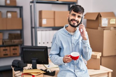 Young hispanic man ecommerce business worker talking on smartphone drinking coffee at office