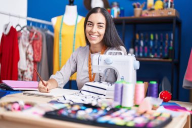 Young beautiful hispanic woman tailor smiling confident drawing on notebook at clothing factory