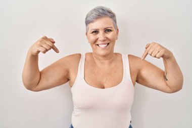 Beyaz arka planda duran orta yaşlı beyaz bir kadın. Yüzünde gülümseme, kendini gururlu ve mutlu olarak gösteren bir kadın.. 