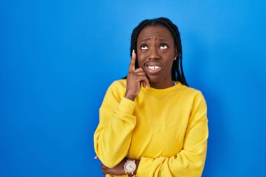 Beautiful black woman standing over blue background thinking worried about a question, concerned and nervous with hand on chin 