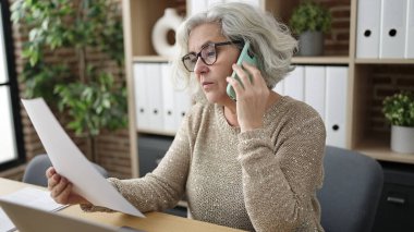 Middle age woman with grey hair business worker talking on smartphone reading document at office