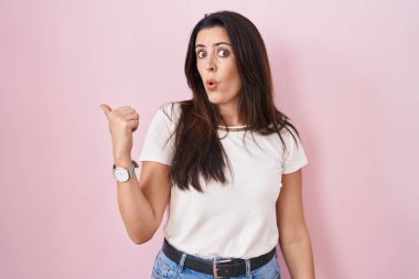 Young brunette woman standing over pink background surprised pointing with hand finger to the side, open mouth amazed expression. 
