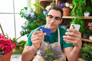 Young hispanic man florist using smartphone holding credit card at flower shop