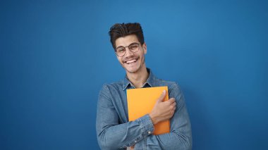 Young hispanic man smiling confident holding book over isolated blue background